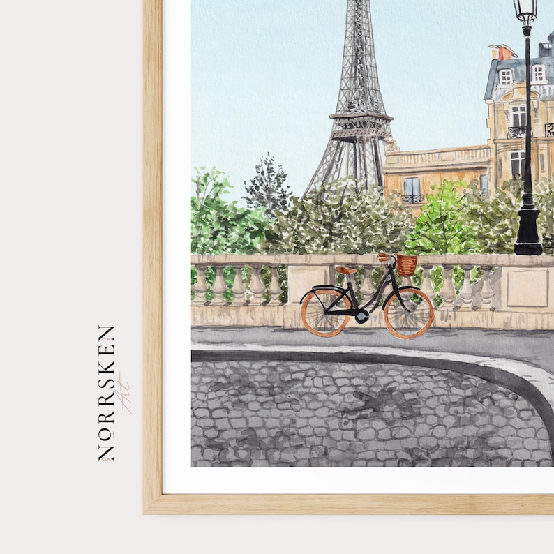 May include: Watercolor print of the Eiffel Tower in Paris, with a bicycle on a cobblestone street. The artwork features a light blue sky and lush green trees, with a decorative stone bridge and buildings in the background. The print is framed in a light wood frame.