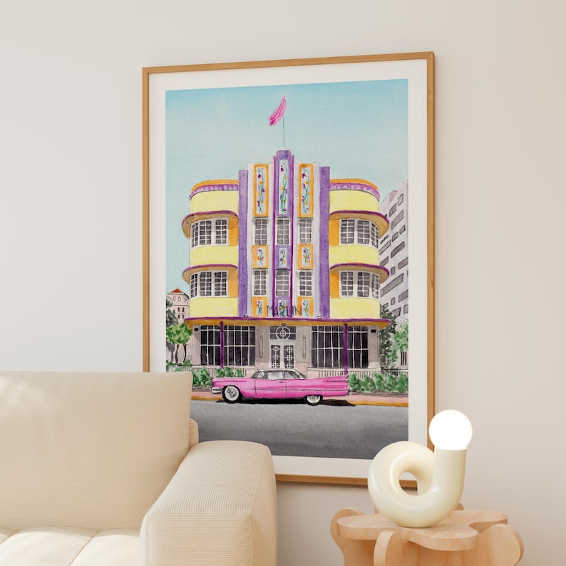 May include: A framed art print depicting a yellow and purple Art Deco building with the word MARLIN on the facade. A long pink vintage car is parked in front of the structure. The scene features a light blue sky and green foliage at the base.