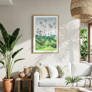 Pode incluir: Uma impress&atilde;o em aquarela emoldurada de uma paisagem com altas palmeiras em uma colina verde sob um c&eacute;u nublado. A obra de arte &eacute; exibida acima de um sof&aacute; branco em uma sala de estar, com uma grande planta em vaso e uma l&acirc;mpada tecida.