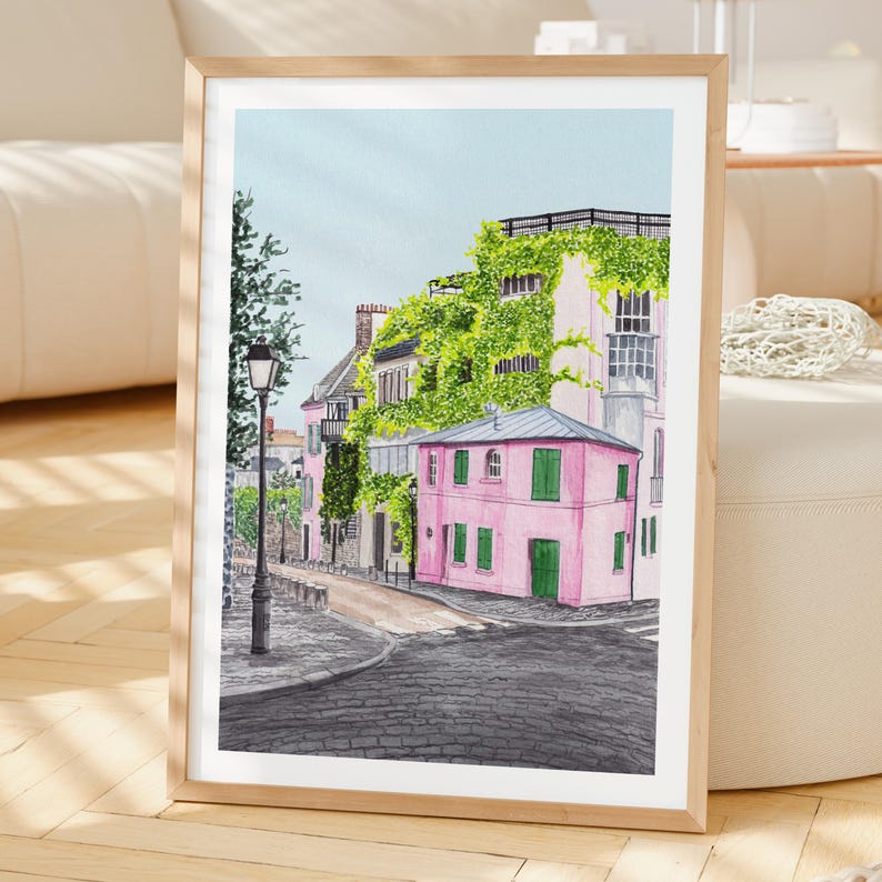 May include: A framed art print featuring a watercolor illustration of a street corner with a pink building covered in green vines, a dark gray cobblestone road, a black street lamp, and a light blue sky, all enclosed in a light wood frame.