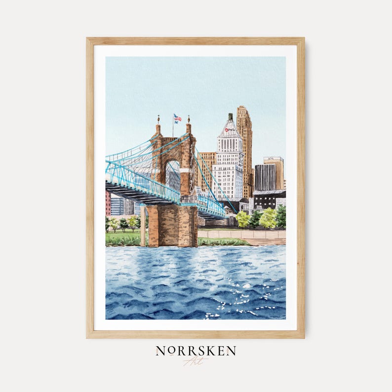 May include: Watercolor painting of a cityscape featuring a blue suspension bridge over a river, with buildings in the background. The artwork is framed in a light wood frame. The water is a deep blue, and the sky is a light blue. The artist's name, NORRSKEN Art, is at the bottom.