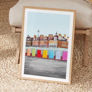May include: A framed art print featuring a row of colorful beach huts in front of multi-story brick buildings under a light blue sky. The beach huts are painted in vertical stripes of orange, green, red, yellow, blue, and pink.