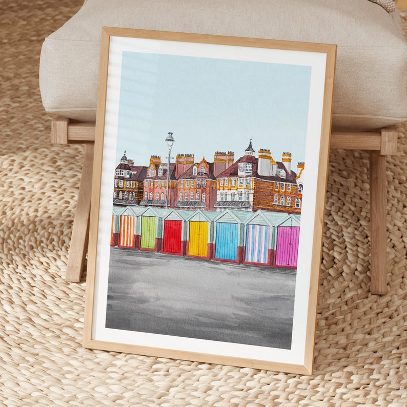 May include: A framed art print featuring a row of colorful beach huts in front of multi-story brick buildings under a light blue sky. The beach huts are painted in vertical stripes of orange, green, red, yellow, blue, and pink.