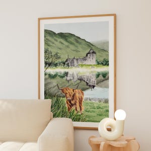 Puede incluir: Impresi&oacute;n en acuarela de una vaca de las Tierras Altas escocesas de pie en hierba alta, con un castillo y monta&ntilde;as reflejadas en el agua. La obra de arte est&aacute; enmarcada en un marco de madera clara y cuelga de una pared neutra.