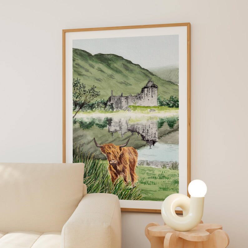 Puede incluir: Impresi&oacute;n en acuarela de una vaca de las Tierras Altas escocesas de pie en hierba alta, con un castillo y monta&ntilde;as reflejadas en el agua. La obra de arte est&aacute; enmarcada en un marco de madera clara y cuelga de una pared neutra.