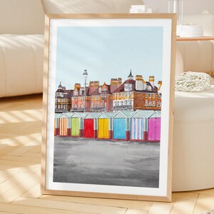 May include: A framed art print featuring a row of colorful beach huts in front of multi-story brick buildings under a pale blue sky. The huts are painted in vertical stripes of orange, green, red, yellow, blue, and pink, set against a gray ground.