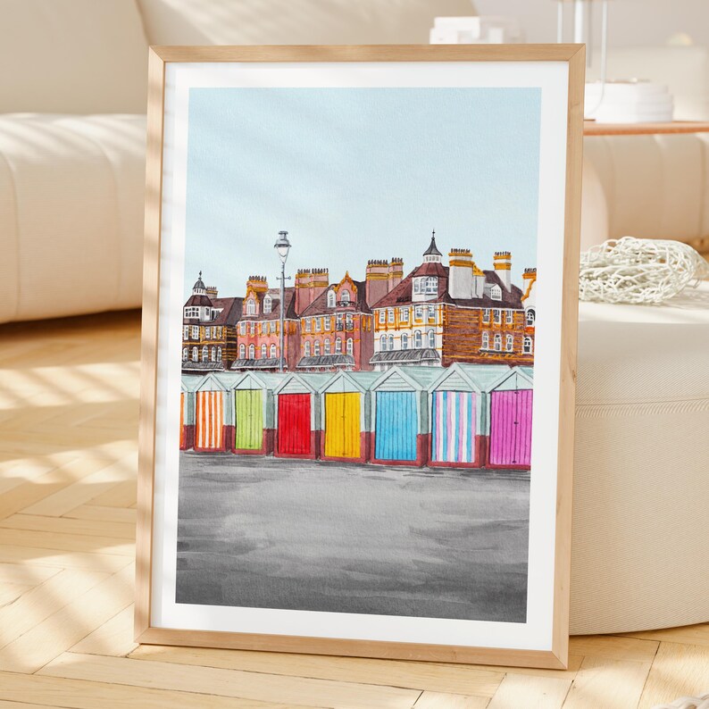 May include: A framed art print featuring a row of colorful beach huts in front of multi-story brick buildings under a pale blue sky. The huts are painted in vertical stripes of orange, green, red, yellow, blue, and pink, set against a gray ground.