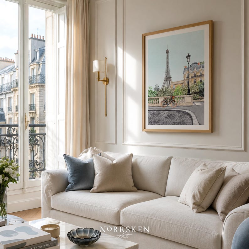 May include: A living room with a cream-colored sofa and throw pillows. A framed print of the Eiffel Tower hangs on the wall. The room has a large window with a view of a city street and buildings. The text "NORRSKEN ART" is visible.