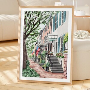 May include: A framed watercolor painting depicting a street scene with historic townhouses, brick sidewalks, and large trees. The buildings feature teal shutters, white porches, and American flags hanging from the facades, with stone steps leading up.