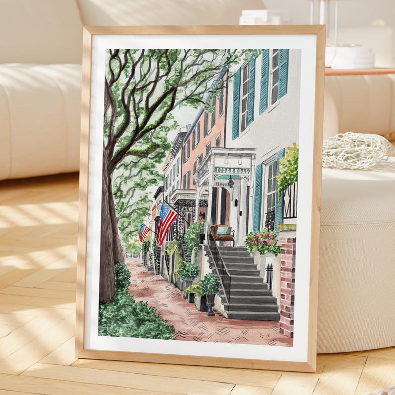 May include: A framed watercolor painting depicting a street scene with historic townhouses, brick sidewalks, and large trees. The buildings feature teal shutters, white porches, and American flags hanging from the facades, with stone steps leading up.