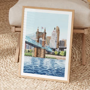May include: Watercolor painting of a cityscape featuring a blue suspension bridge and tall buildings. The artwork is framed in a light wood frame and depicts a river with rippling water under a light blue sky. The artwork is displayed on a woven surface.