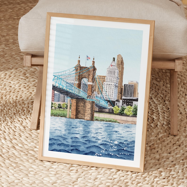 May include: Watercolor painting of a cityscape featuring a blue suspension bridge and tall buildings. The artwork is framed in a light wood frame and depicts a river with rippling water under a light blue sky. The artwork is displayed on a woven surface.