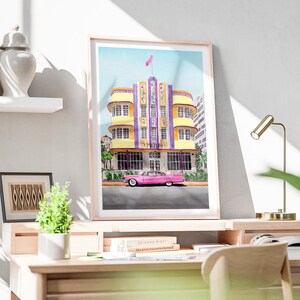 May include: A framed watercolor print depicting a yellow and purple Art Deco building with a pink vintage car parked in front. The building features vertical panels, multiple windows, and a pink flag on the roof. The artwork is displayed on a desk.