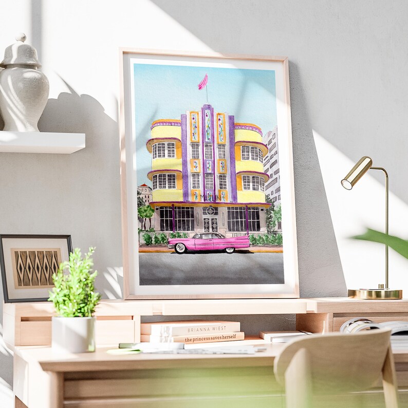 May include: A framed watercolor print depicting a yellow and purple Art Deco building with a pink vintage car parked in front. The building features vertical panels, multiple windows, and a pink flag on the roof. The artwork is displayed on a desk.