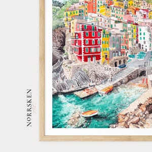 May include: A watercolour print in a light wood frame depicting a coastal village with stacked red, yellow, and white buildings on a rocky cliffside overlooking turquoise water with small boats. The text NORRSKEN Art is printed on the left side.