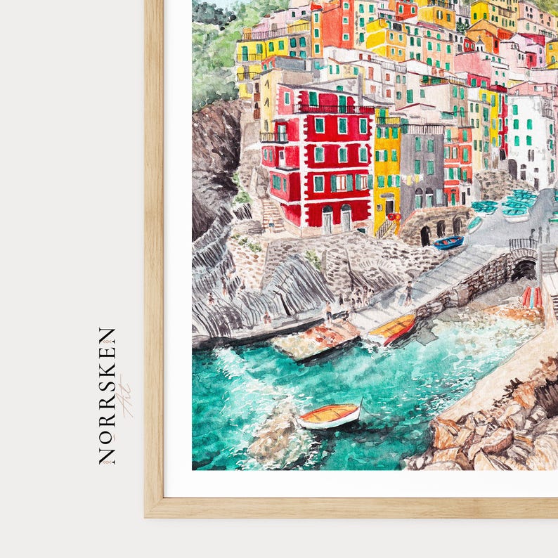 May include: A watercolour print in a light wood frame depicting a coastal village with stacked red, yellow, and white buildings on a rocky cliffside overlooking turquoise water with small boats. The text NORRSKEN Art is printed on the left side.