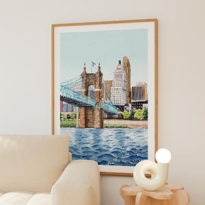 May include: Watercolor painting of a cityscape featuring a blue suspension bridge with a brown stone structure. The bridge spans over blue water, with buildings and trees in the background. The artwork is framed in a light brown wooden frame.