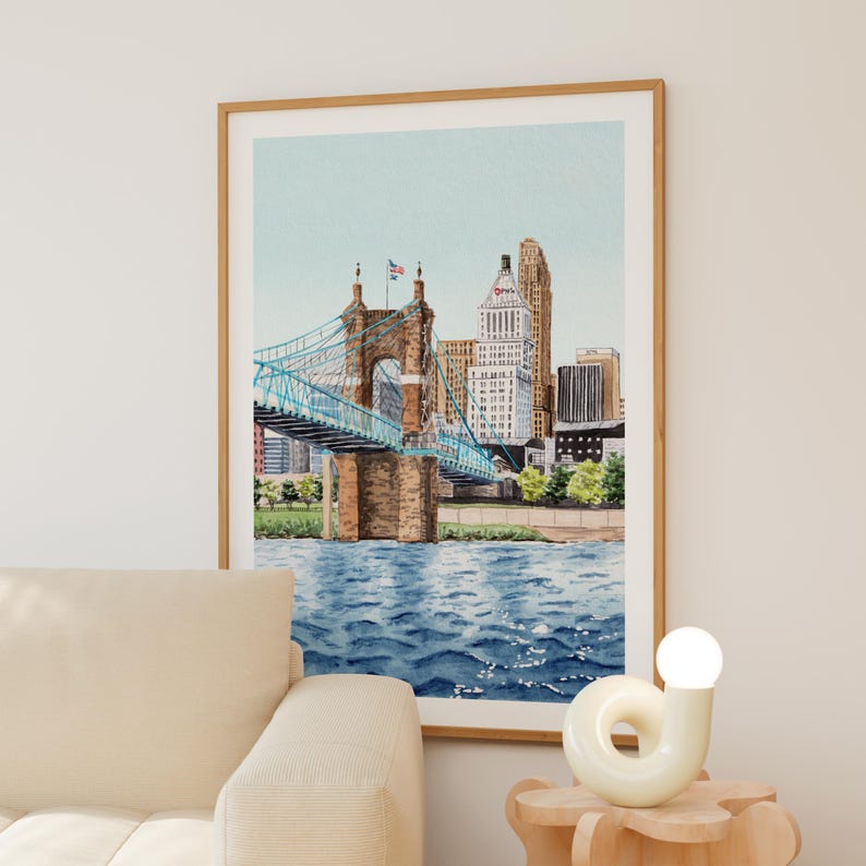 May include: Watercolor painting of a cityscape featuring a blue suspension bridge with a brown stone structure. The bridge spans over blue water, with buildings and trees in the background. The artwork is framed in a light brown wooden frame.