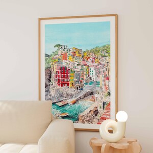 May include: A framed watercolour print depicting a coastal village with colourful houses in shades of red, yellow, and white, built into a rocky cliffside overlooking a turquoise harbour with small boats and a stone pier under a clear blue sky.