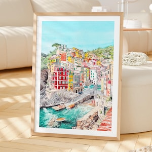 May include: A framed watercolour painting depicting a coastal village with colourful houses stacked on a rocky cliffside overlooking a turquoise harbour with small boats, all set against a clear blue sky in a vertical rectangular orientation.