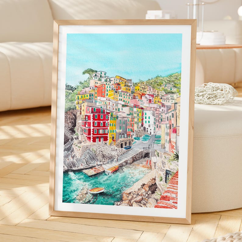 May include: A framed watercolour painting depicting a coastal village with colourful houses stacked on a rocky cliffside overlooking a turquoise harbour with small boats, all set against a clear blue sky in a vertical rectangular orientation.