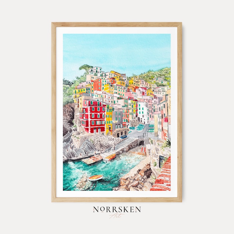 May include: A framed watercolour print depicting a coastal village with colourful red, yellow, and white buildings nestled on a rocky cliffside above turquoise water. A small boat floats in the harbour, and the text NORRSKEN Art appears below.