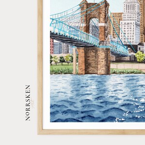 May include: Watercolor painting of a suspension bridge with blue accents, spanning a river. The bridge is brown with a blue deck and supports. City buildings are in the background. The artwork is framed in a light wood frame. The text "NORRSKEN" is on the left side.