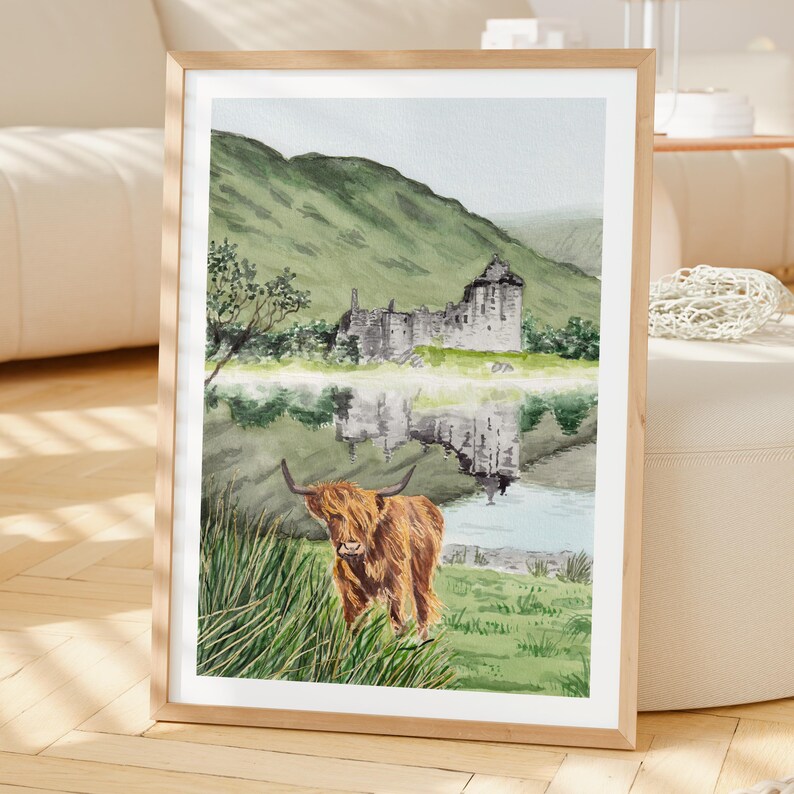 Puede incluir: Pintura de acuarela enmarcada de una vaca de las Tierras Altas en la hierba, con una ruina de castillo y una monta&ntilde;a al fondo. La escena se refleja en el agua. El marco es de madera clara.