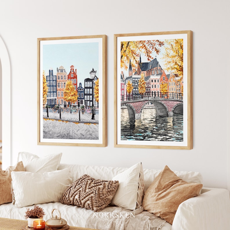 May include: Two framed watercolour prints of European cityscapes. One shows colourful buildings along a canal with bicycles, the other a bridge over water. Both feature autumn foliage and are displayed above a white sofa with cushions.