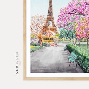 May include: A watercolor print featuring the Eiffel Tower in the background, a carousel, and a park path with a green bench. The text NORRSKEN Art is printed vertically on the left side. The scene includes trees with pink and orange foliage.