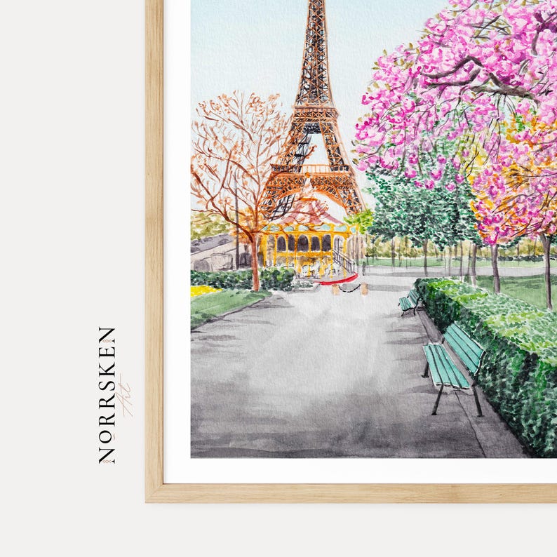 May include: A watercolor print featuring the Eiffel Tower in the background, a carousel, and a park path with a green bench. The text NORRSKEN Art is printed vertically on the left side. The scene includes trees with pink and orange foliage.