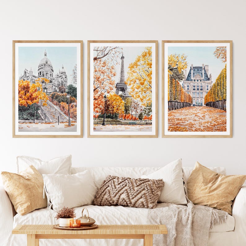K&ouml;nnte beinhalten: Drei gerahmte Aquarelldrucke von Pariser Wahrzeichen: Sacr&eacute;-C&oelig;ur-Basilika, Eiffelturm und ein gro&szlig;es Geb&auml;ude mit einem von B&auml;umen ges&auml;umten Weg. Das Kunstwerk zeigt Herbstfarben mit orangefarbenem und gelbem Laub. Die Drucke werden &uuml;ber einem wei&szlig;en Sofa mit dekorativen Kissen ausgestellt.