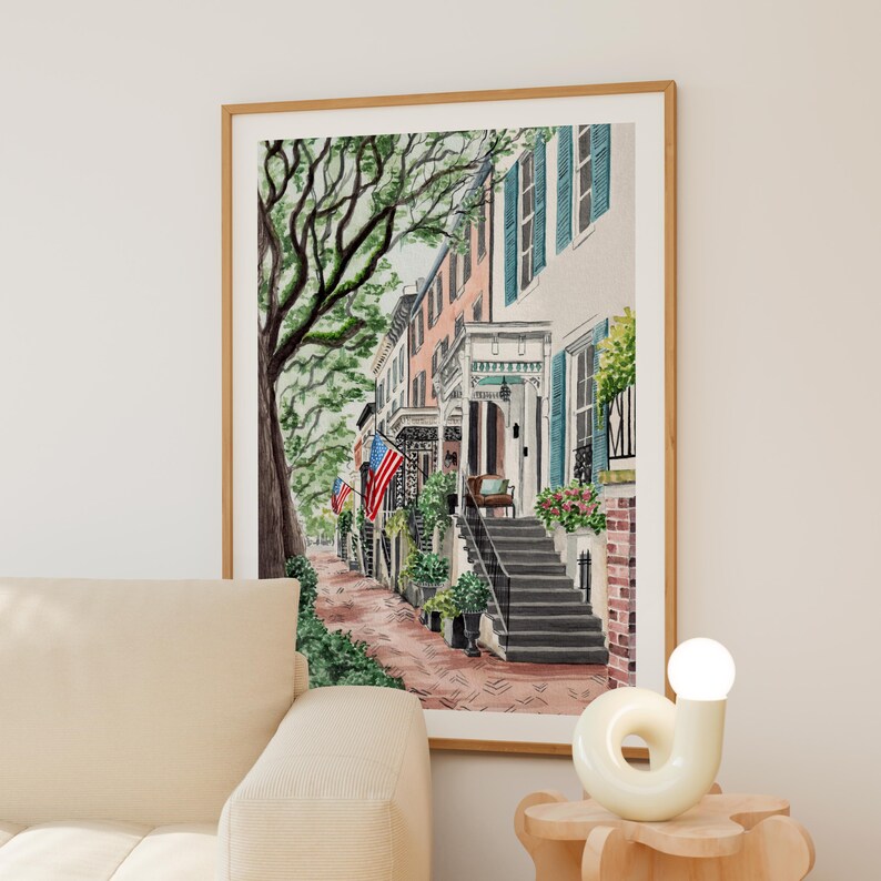 May include: A framed watercolor print depicting a row of historic townhouses with teal shutters, brick steps, and American flags. A large tree with green foliage stands on the left side of the street, while a brick walkway leads toward the houses.