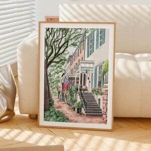 May include: A framed watercolor painting depicting a street scene with historic townhouses, a large tree, and American flags. The architecture features light-colored facades, blue shutters, and stone steps leading to porches, all set along a brick walkway.