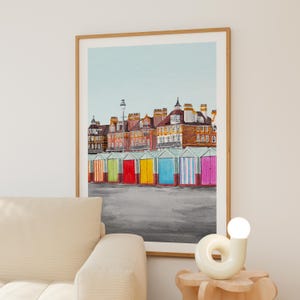 May include: A framed watercolor print depicting a row of colorful beach huts in front of multi-story brick buildings under a pale blue sky. The huts are painted in vibrant shades of orange, green, red, yellow, blue, and pink against a gray ground.