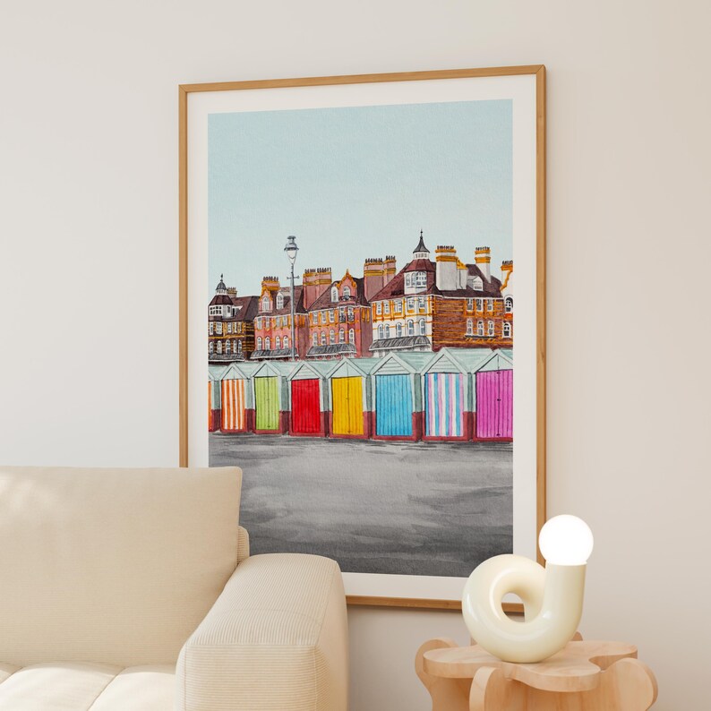 May include: A framed watercolor print depicting a row of colorful beach huts in front of multi-story brick buildings under a pale blue sky. The huts are painted in vibrant shades of orange, green, red, yellow, blue, and pink against a gray ground.