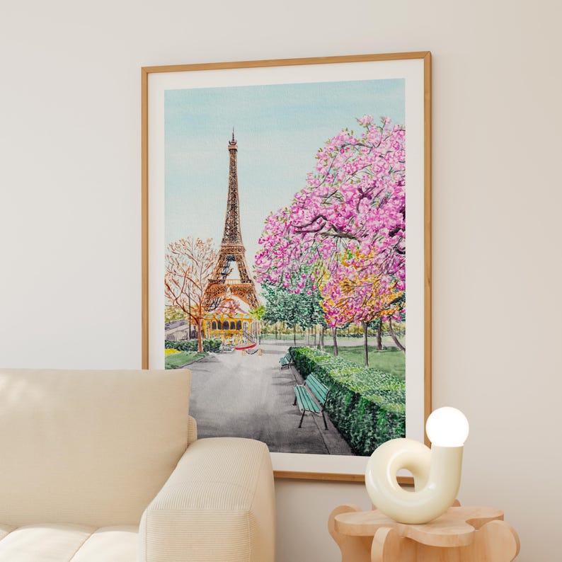May include: A framed watercolor print depicting the Eiffel Tower in Paris, surrounded by vibrant pink cherry blossom trees and green hedges. A park pathway leads toward the tower, featuring a green bench and a clear, light blue sky above.