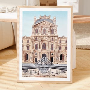 K&ouml;nnte beinhalten: Gerahmtes Aquarellgem&auml;lde des Louvre-Museums in Paris. Das Kunstwerk zeigt das ber&uuml;hmte Geb&auml;ude mit seiner detaillierten Architektur, einer Glaspyramide und einer Spiegelung im Wasser, vor blauem Himmel. Der Rahmen ist aus hellem Holz.