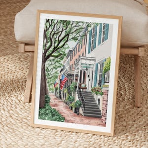 May include: A framed watercolor print depicting a street scene with historic townhouses, a large tree, and American flags. The architecture features brick stairs, blue shutters, and a pink facade, all rendered in a soft, artistic style.