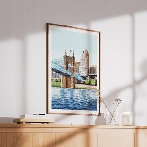 May include: Watercolor print of a bridge with a blue suspension design over a river, with a city skyline in the background. The artwork is framed in a wooden frame and displayed on a wooden cabinet.