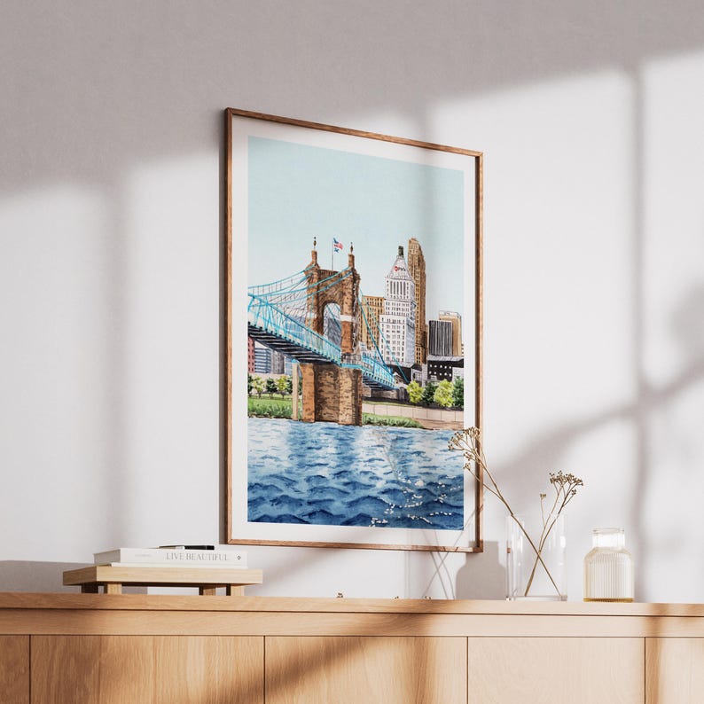 May include: Watercolor print of a bridge with a blue suspension design over a river, with a city skyline in the background. The artwork is framed in a wooden frame and displayed on a wooden cabinet.