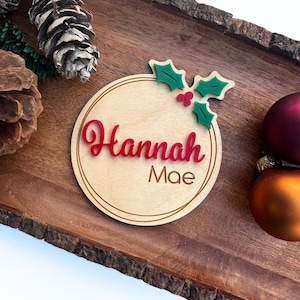 May include: A round wooden ornament with a holly sprig and red berries. The ornament is personalized with the name "Hannah Mae" in red lettering.