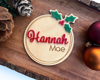 May include: A round wooden ornament with a holly sprig and red berries. The ornament is personalized with the name "Hannah Mae" in red lettering.