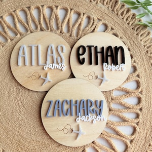May include: Three wooden round name plaques with names in various colors. The names are Atlas, Ethan, and Zachary, with additional names below. Each plaque has a small airplane graphic and dotted flight path. The background is a woven, tan surface.