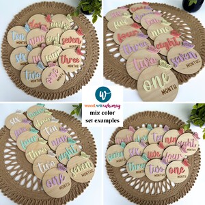 May include: A set of 12 wooden baby milestone discs with pastel pink, yellow, and green butterflies. The discs are numbered one through twelve and have the word "month" written on each disc. The discs are arranged in a circle on a woven mat.