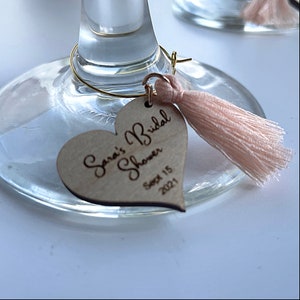 May include: A wooden heart-shaped tag with the text "Sara's Bridal Shower Sept 15 2021" engraved on it. The tag is attached to a glass with a gold wire and a pink tassel.