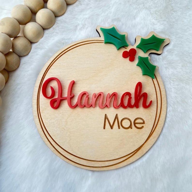 May include: A round wooden ornament with a red and white name "Hannah Mae" and a green holly sprig with red berries.