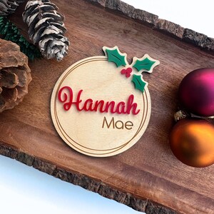 May include: A round wooden ornament with a red holly design and the name "Hannah Mae" written in red cursive lettering.