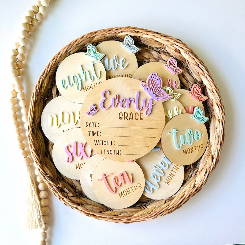 May include: A set of 10 wooden baby milestone discs with pastel coloured butterflies and the words "eight months", "nine months", "ten months", "eleven months", "two months", "six months", "we months", "Evely Grace" with spaces for date, time, weight, and length.