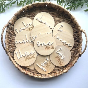 May include: A set of twelve wooden baby milestone discs in a wicker basket. Each disc features a white number and the word "month" in a cursive font. The numbers range from one to twelve.